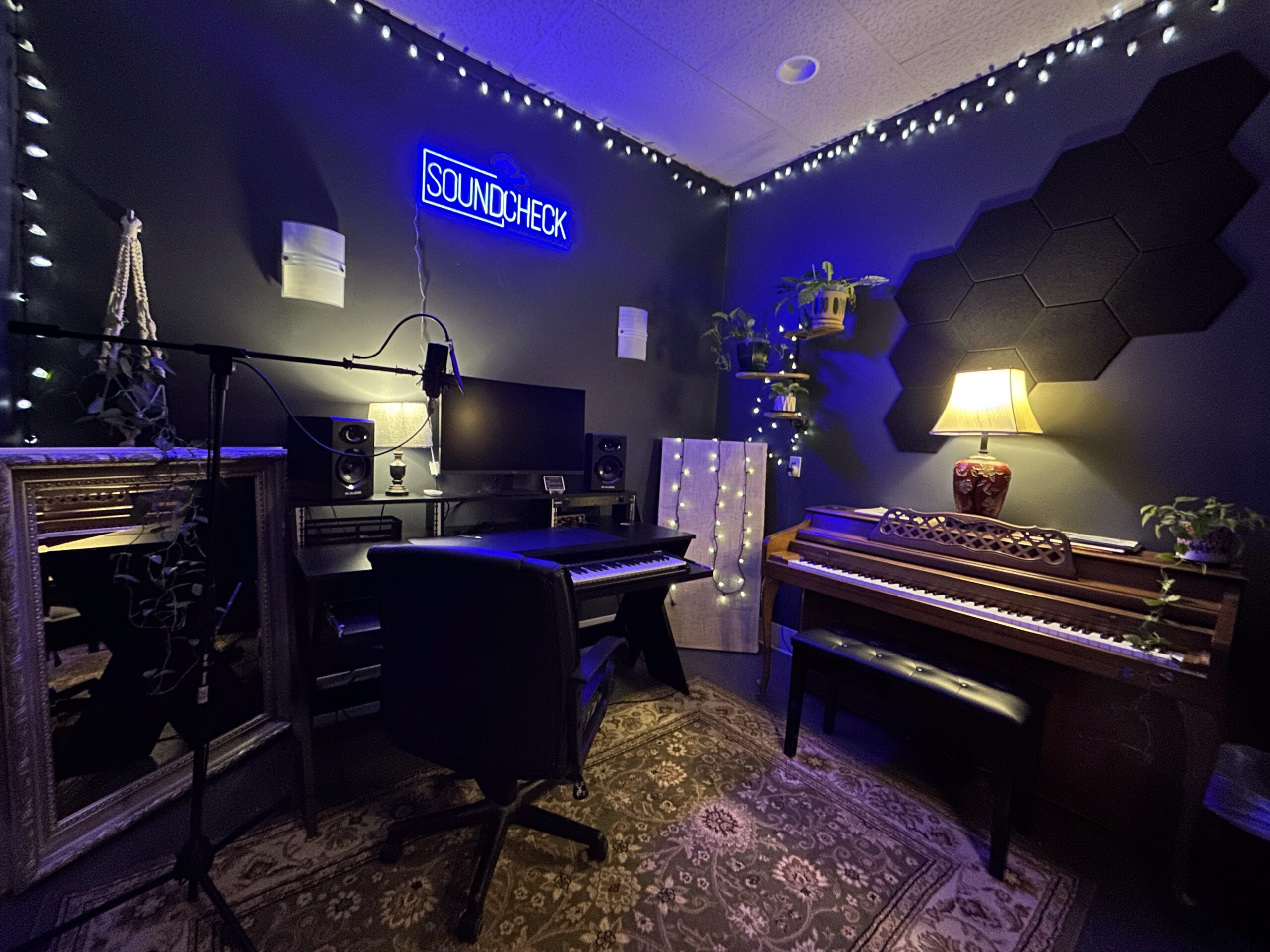The recording room bathed in blue light - studio desk with monitors and a MIDI keyboard, the SOUNDCHECK neon sign, hexagon acoustic panels, and a vintage upright piano